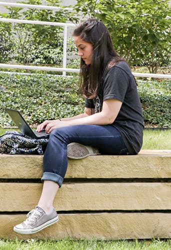 A student sits outdoors on a ledge with a laptop